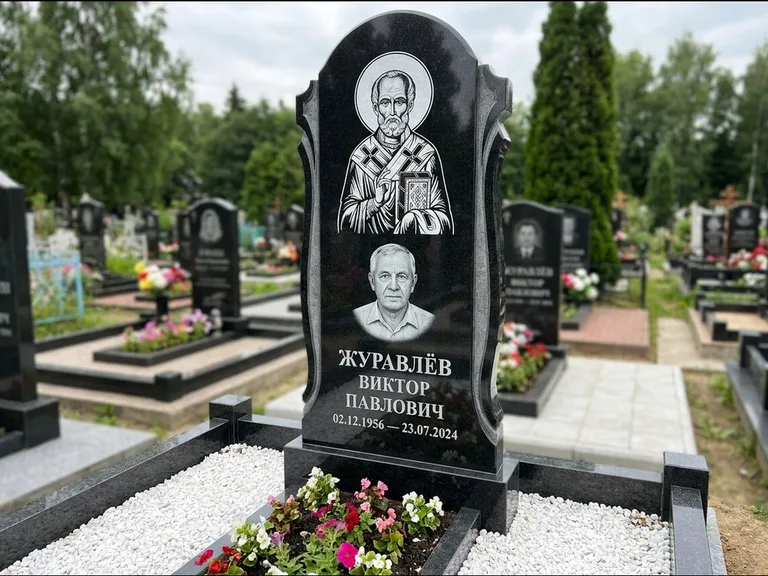 Памятник с гравировкой иконы: стела с образом в полный рост на обороте — гравировка образа Матроны Московской, низкие кусты спиреи в цвету, работа «Лебедева» в Новосибирске